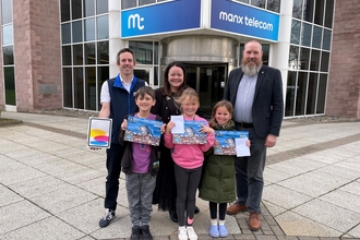 Three happy kids holding a wildlife calendar with a seal and three adults standing behind holding an iPad in front of the Manx Telecom building.