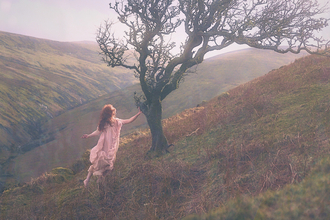 A woman wearing a dress is posing by a windswept tree on a hillside