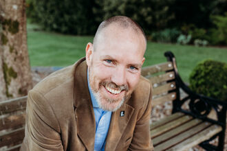Graham sat on a park bench under a tree looking up at the camera smiling