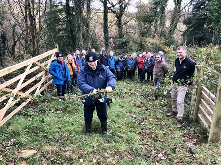 The picture shows His Excellecny, Sir john lorimer, cutting a 'ribbon' of ive across a gateway with trees behind and a group of other people in outdoor gear.