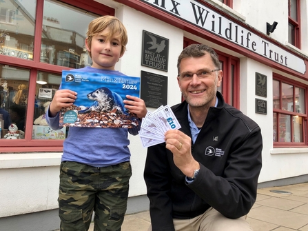 Small boy smiling holding a wildlife calendar next to man wearing a black MWT coat and brown hair. 