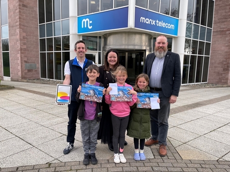 Three happy kids holding a wildlife calendar with a seal and three adults standing behind holding an iPad in front of the Manx Telecom building.