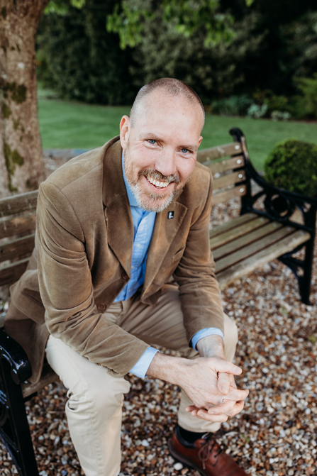 Graham sat on a park bench under a tree looking up at the camera smiling