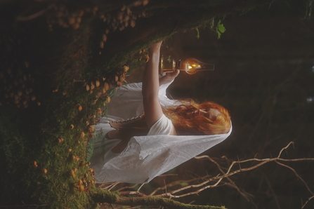 A woman holds a lantern in the woods, she is stood by a moss covered tree that is sprouting mushrooms.