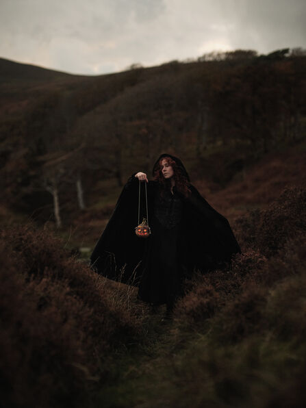 A woman dressed in a black cloak holding a candle lit turnip in the uplands