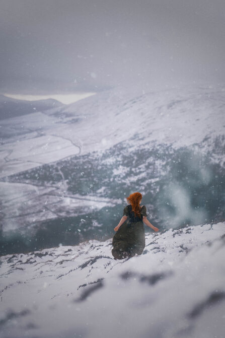 A woman in a black dress stands on the snowy hillside looking off into the distance.