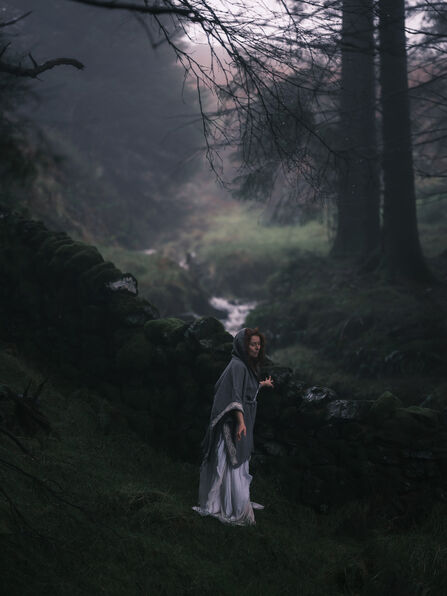 A woman with a Celtic cloak roams the woods, she is stood by a stream.
