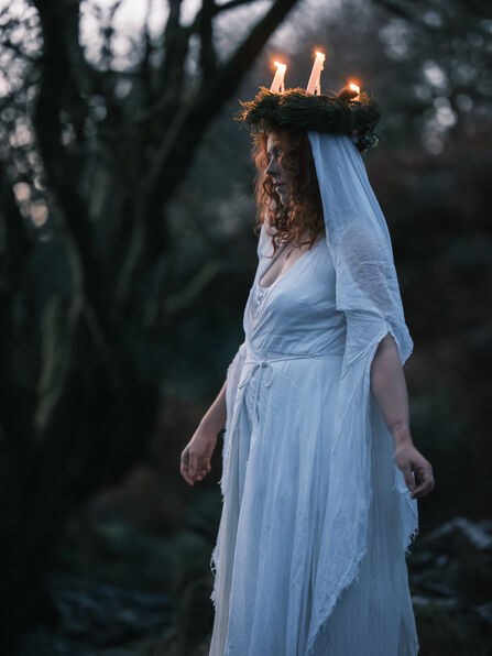 A woman in a white dress wearing a candle lit crown wreath in the woods.