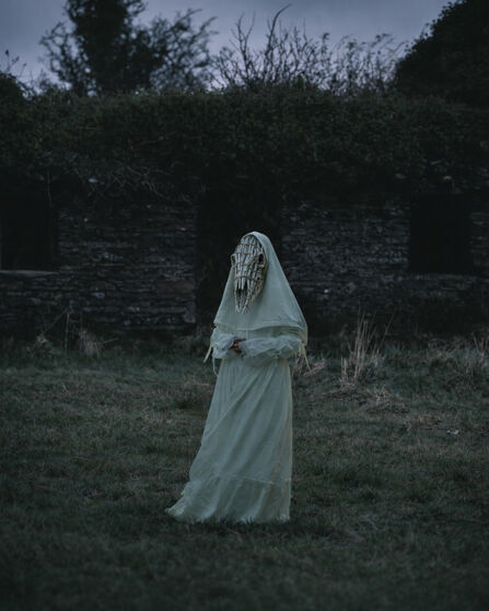 A women in a white dress wears a horse skull mask made of sticks, she is stood in front of an old tholtan.