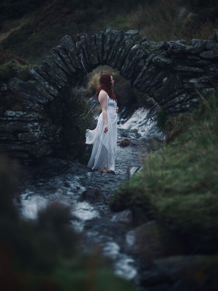 A woman in a white dress with red hair stand under a stone bridge in the moorlands.
