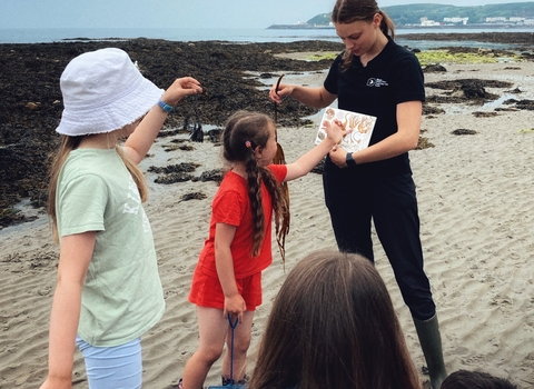 Education Office Beth holding a piece of seaweed and an ID guide with a group of children.