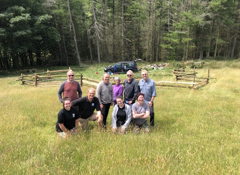 A work party of 9 people on a grassy hill sloping down towards trees behind and a wooden hand made appalachian fence